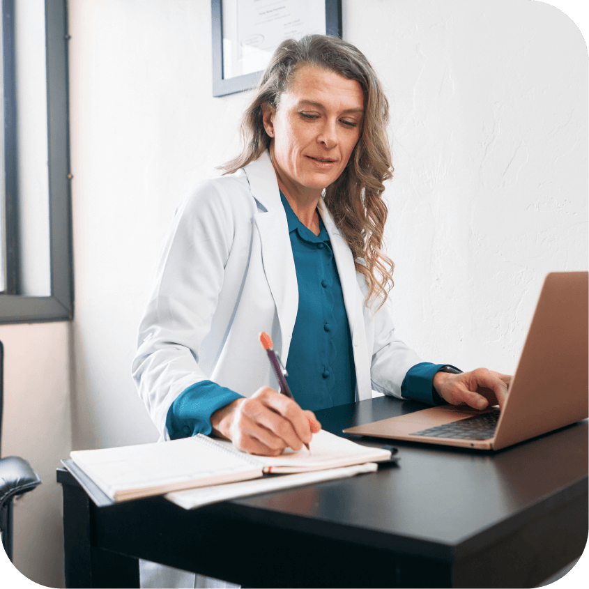 A person in a doctors coat, sits at a wood desk, and looks down at a note pad while taking notes next to a computer.