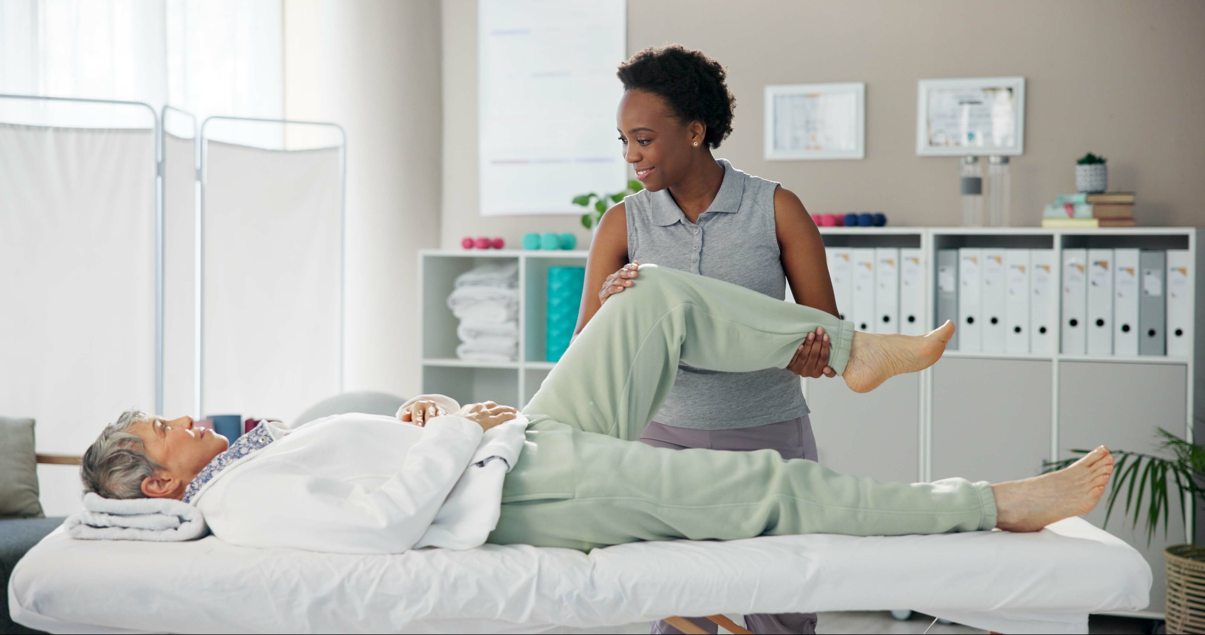 A woman lies on a treatment bed in a physical therapy clinic while her physical therapist helps her perform a stretch for her leg and back.